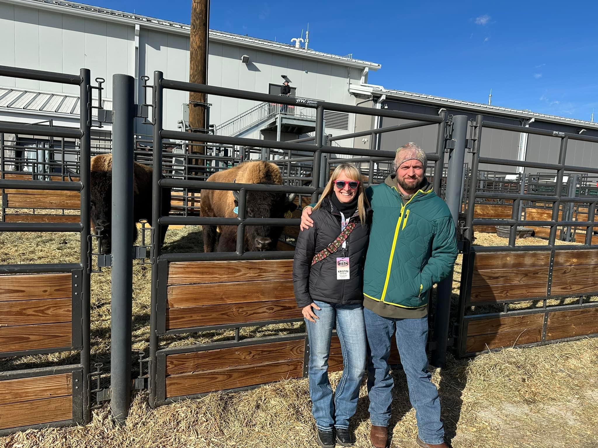 A woman and a man stand together in front of a bison enclosure at a livestock event, with bison visible in the background. The setting features metal fencing and a clear blue sky, indicating a sunny day.