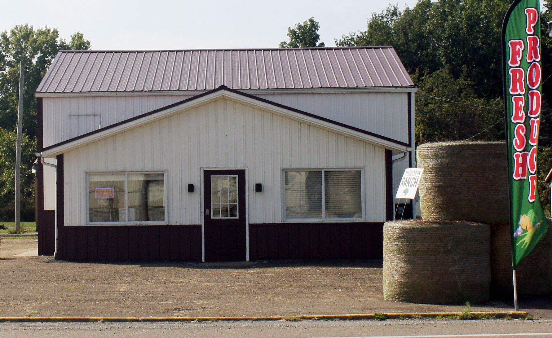 Small rural produce shop with a white and brown exterior, featuring an "Open" sign in the window, and hay bales nearby, promoting fresh local products.